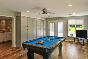 Une table de billard avec des billes, un ventilateur de plafond et un téléviseur dans une piÚce au plancher en bois et aux murs de couleur claire.