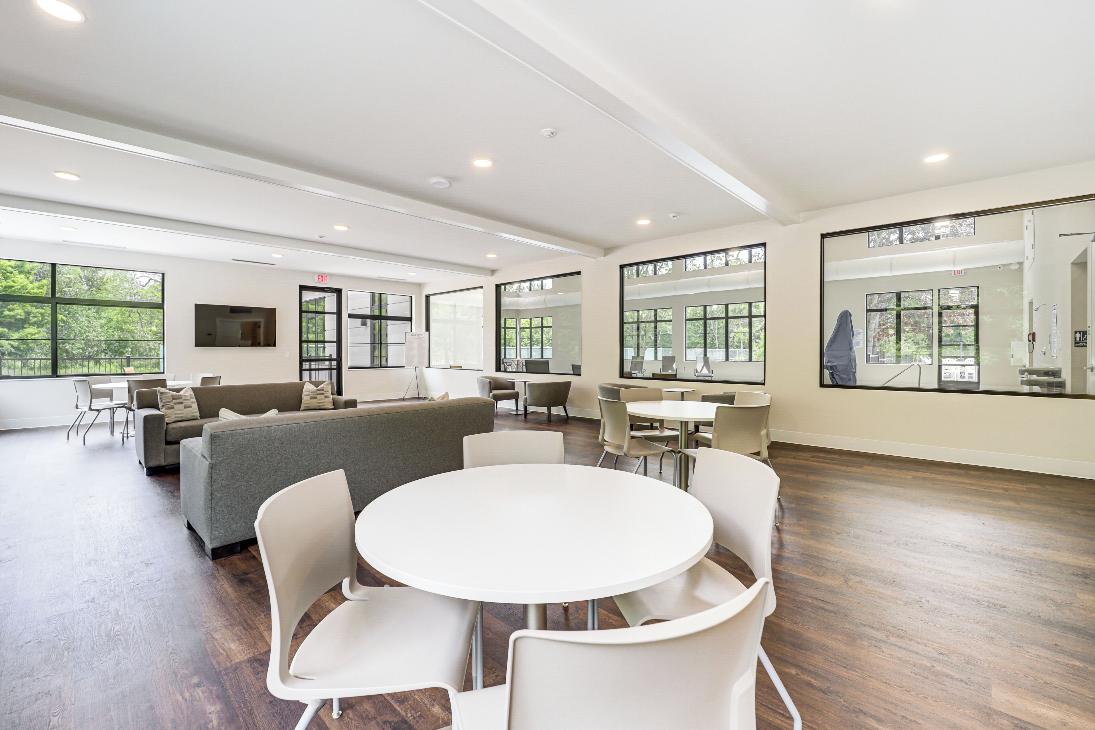 A spacious, modern room with a large round table and white chairs, a grey sofa, and a TV mounted on the wall.