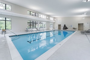 A large indoor swimming pool with multiple lanes, sunlit by natural light from large windows, and equipped with lounge chairs along the sides.