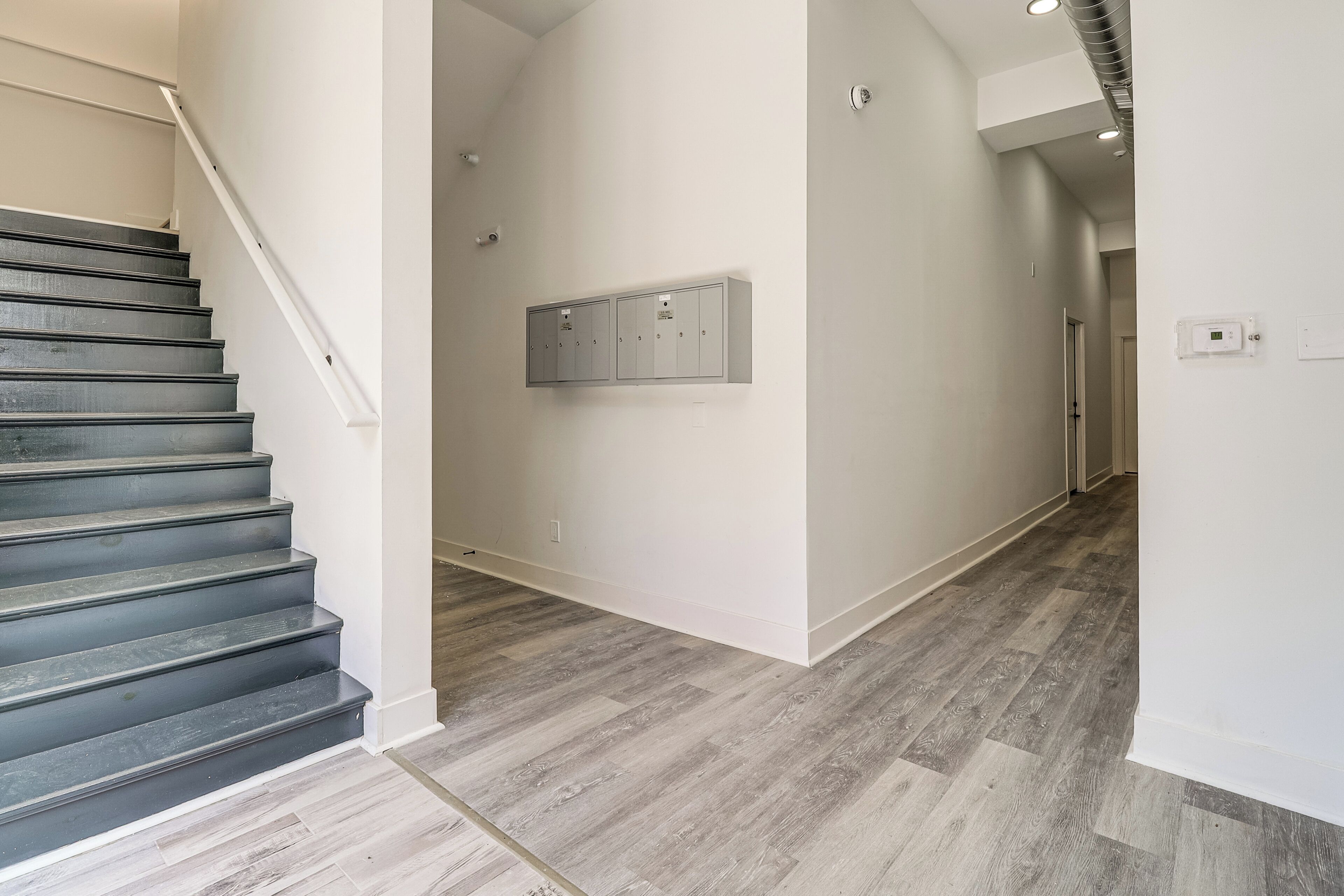 Una escalera conduce a un pasillo con piso de madera, un tablero eléctrico en la pared y una puerta al final.