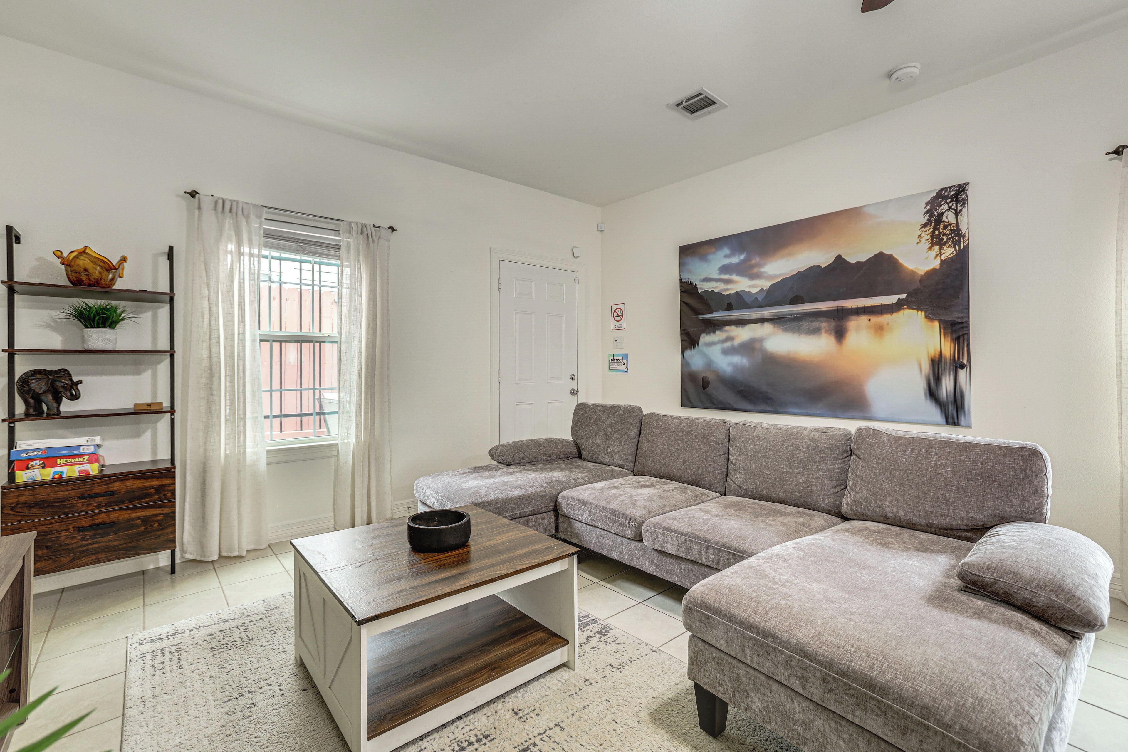 A living room with a grey sofa, a coffee table, a bookshelf, a painting, and a window with curtains.