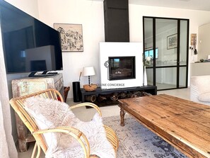 A modern living room with a fireplace, a television, wicker furniture, and a glass door leading to a balcony. - Spacious 5-bedroom house with AC and Pool (Le Plan-de-la-Tour)