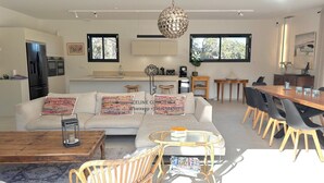 A modern living room with a sofa, coffee table, wicker chairs, a dining table with chairs, a hanging light fixture, and a kitchen area with cabinets and a refrigerator. - Spacious 5-bedroom house with AC and Pool (Le Plan-de-la-Tour)