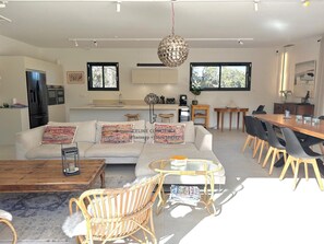Een moderne woonkamer met een bank, salontafel, rieten stoelen, een eettafel met stoelen, een hanglamp en een keuken met kasten en een koelkast.