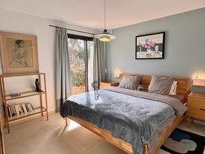 A bedroom with a bed, a nightstand, a shelf with books, a painting, and a window with curtains. - Spacious 5-bedroom house with AC and Pool (Le Plan-de-la-Tour)