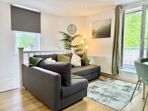 A modern living room with a grey sofa, a potted plant, a floor lamp, and a view of greenery through the window. - Stylish 2-bed Bristol Flat w Roof Garden & Parking (Bristol)