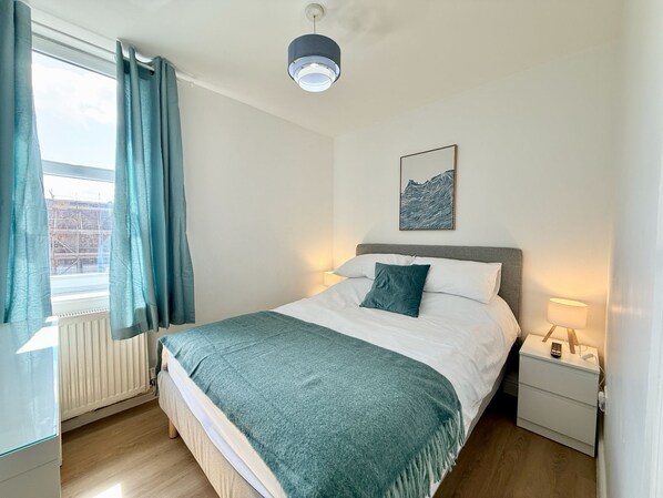 Een slaapkamer met een bed, nachtkastje, lamp, raam met gordijnen en een kunstwerk aan de muur.