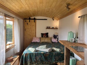 A cozy bedroom with a wooden ceiling, a bed with floral bedding, a bedside table with a kettle, and a window with curtains. - Shepherd Hut (Gloucester)