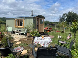A small wooden cabin with a window, a table with a floral tablecloth, and outdoor seating. - Shepherd Hut (Gloucester)