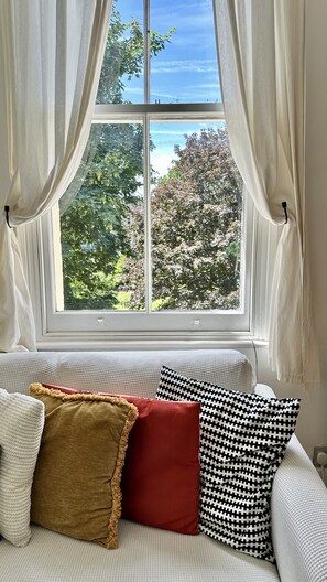 Ein Fenster mit Vorhängen, Blick auf Bäume und ein Sofa mit Kissen.