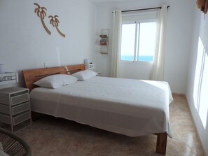 Ein ordentlich eingerichtetes Schlafzimmer mit einem hölzernen Kopfteil, einem Bett mit weißer Bettwäsche, einem Nachttisch, einer kleinen Bücherablage und Meerblick durch ein Fenster.