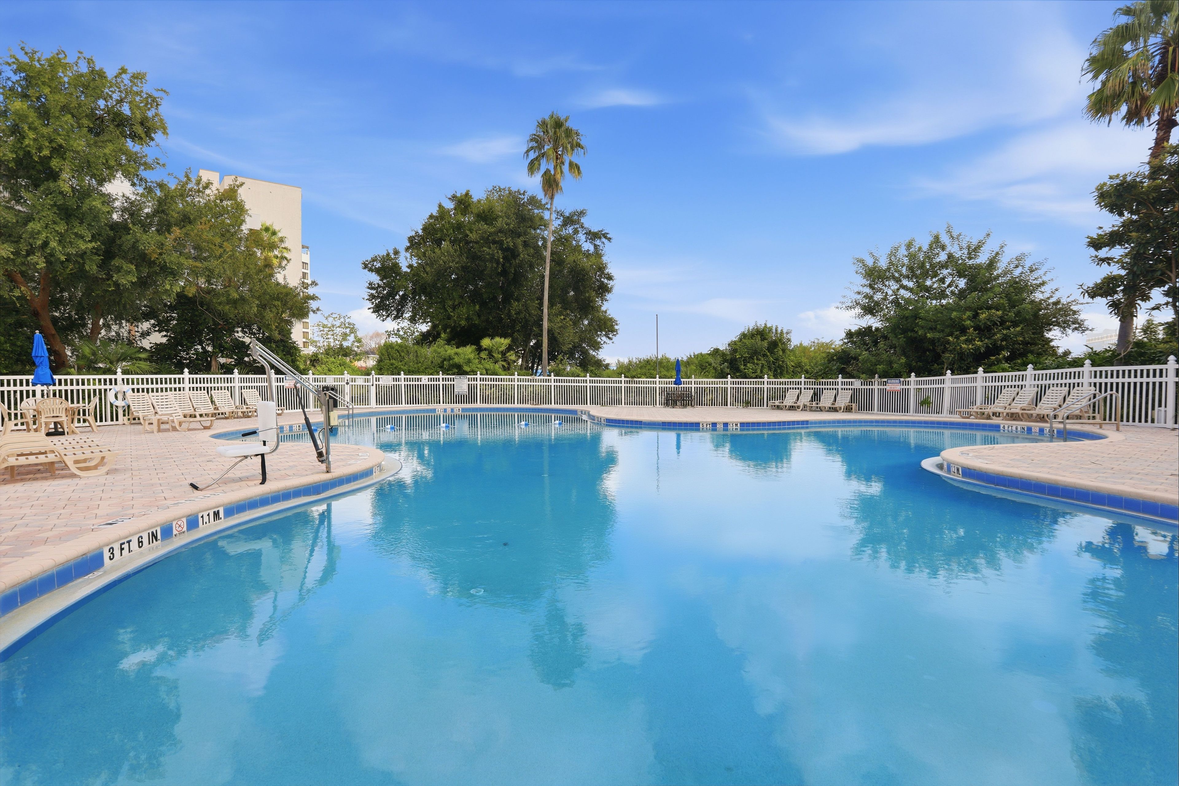 Une piscine entourée d’une clôture blanche, avec des chaises longues et des arbres en arrière-plan.