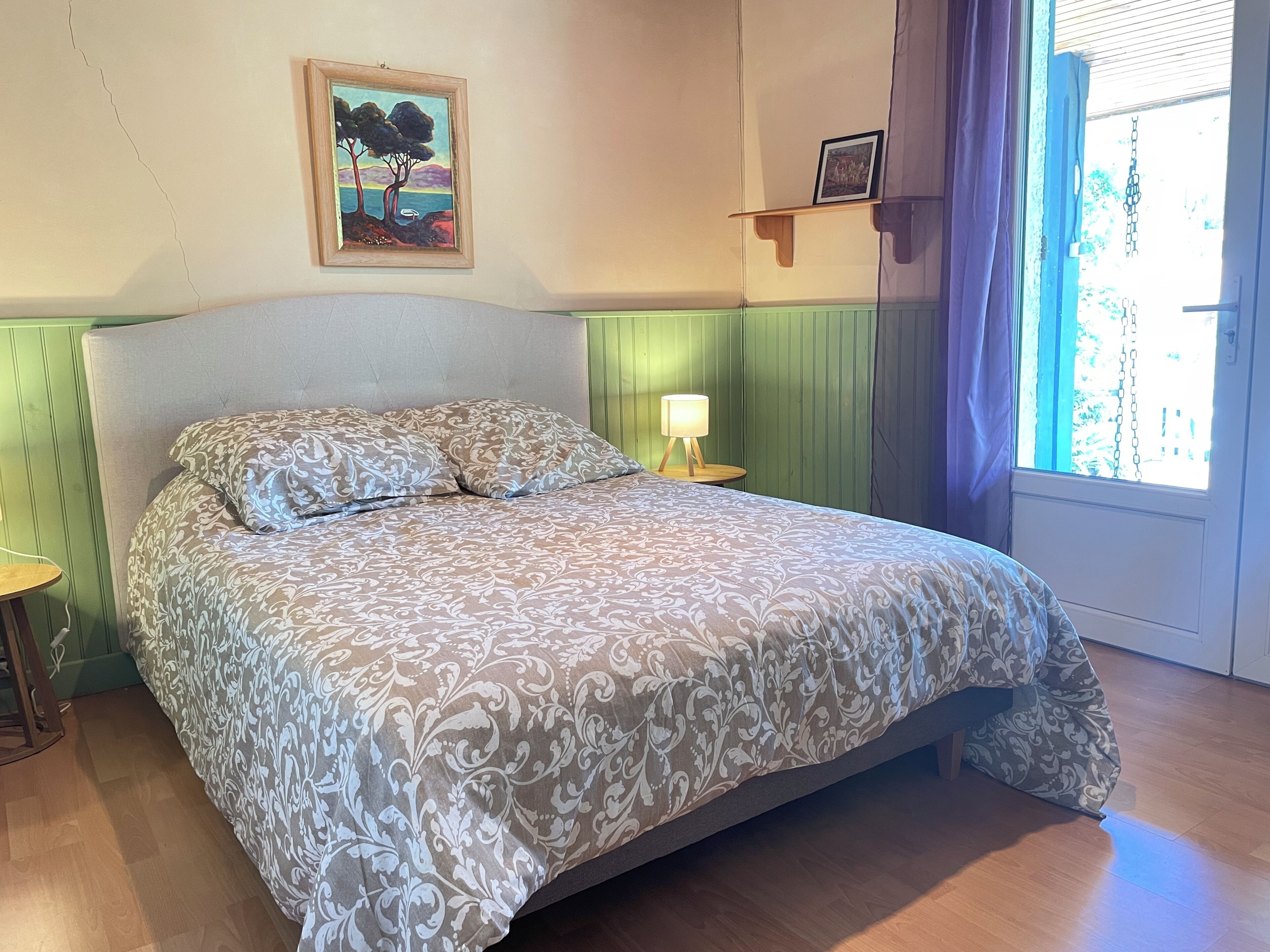A bedroom with a bed, a nightstand, a painting, a shelf with a picture, and a window with curtains.