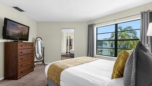 Quarto de hotel com uma cama grande, cômoda de madeira, televisão de tela plana, espelho e janela com vista para palmeiras.