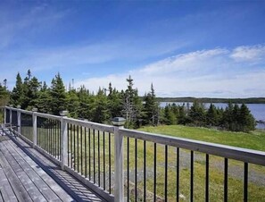 A wooden deck with a railing overlooking a lake and forest. - Rhodies Pondside<br>It's ideal for guests looking to unwind, enjoy scenic views, and experience a quiet getaway. (Placentia Junction)