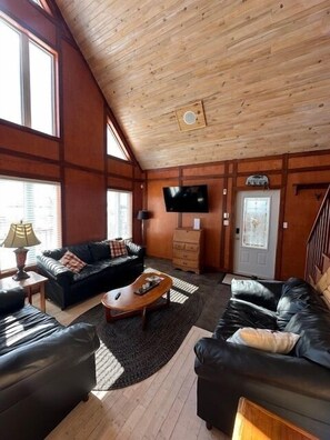 A living room with a wooden ceiling, a television, a sofa, a coffee table, and a fireplace. - Rhodies Pondside<br>It's ideal for guests looking to unwind, enjoy scenic views, and experience a quiet getaway. (Placentia Junction)