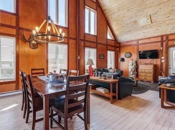 Un salón espacioso con techo de madera, una zona de comedor con mesa y sillas de madera, y un televisor montado en la pared.