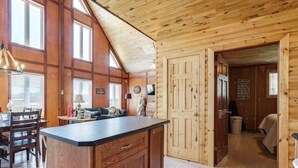 Interior de una cabaña de madera con una isla de cocina, comedor y una puerta visible que da a otra habitación.