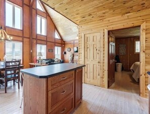 A wooden cabin interior with a kitchen island, dining area, and a visible door leading to another room. - Rhodies Pondside<br>It's ideal for guests looking to unwind, enjoy scenic views, and experience a quiet getaway. (Placentia Junction)
