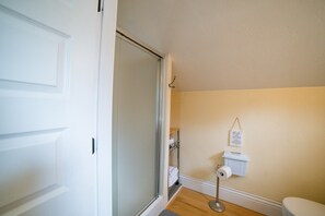 A bathroom with a toilet, a roll of toilet paper, a shelf with towels, and a shower door. - Hayward Field Studio. In the heart of Tracktown USA. (Eugene)