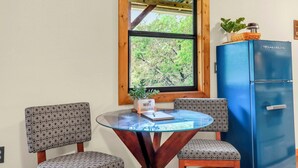 Ein Zimmer mit einem runden Glastisch, zwei gemusterten Stühlen, einem blauen Kühlschrank, einem Fenster mit Blick auf Bäume und einer kleinen Topfpflanze auf dem Kühlschrank.