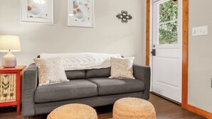Ein graues Sofa mit weißer Decke, zwei gerahmte abstrakte Gemälde, ein dekoratives Metallobjekt und eine Holztür mit Fenster.