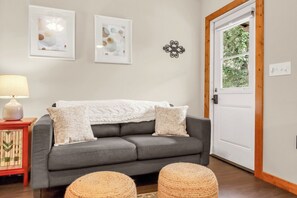 Ein graues Sofa mit weißer Decke, zwei gerahmte abstrakte Gemälde, ein dekoratives Metallobjekt und eine Holztür mit Fenster.