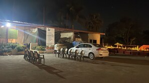 A hotel with a covered outdoor seating area, a white car parked nearby, and a sign listing amenities like rooms, swimming pool, and restaurant. - Buta Inn and Resorts (Vasai)