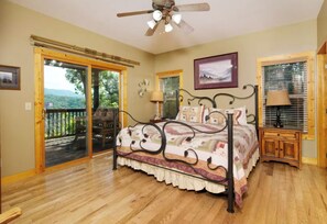 A bedroom with a bed, a ceiling fan, a nightstand, a lamp, a window with blinds, and a door leading to an outdoor deck. - Riversong Retreat | Pet-Friendly Gatlinburg Cabin with Mountain Views & Game Roo (Sevierville)