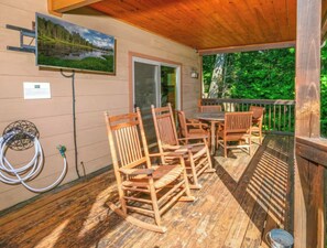 A wooden deck with a table and chairs, a hose, and a painting on the wall. - Riversong Retreat | Pet-Friendly Gatlinburg Cabin with Mountain Views & Game Roo (Sevierville)