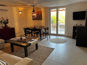 Ein Wohnzimmer mit Sofa, Couchtisch, Fernseher, Essbereich mit Stühlen und einer Glastür zum Außenbereich.