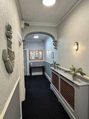A hallway with a mirror, a light fixture, a potted plant, and a door leading to a room.