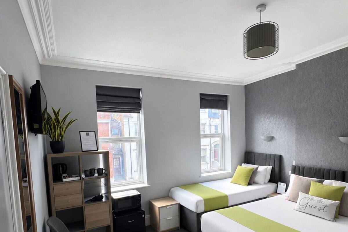 A modern hotel room with a large bed, two bedside tables, a desk with a chair, a small side table, and a potted plant.
