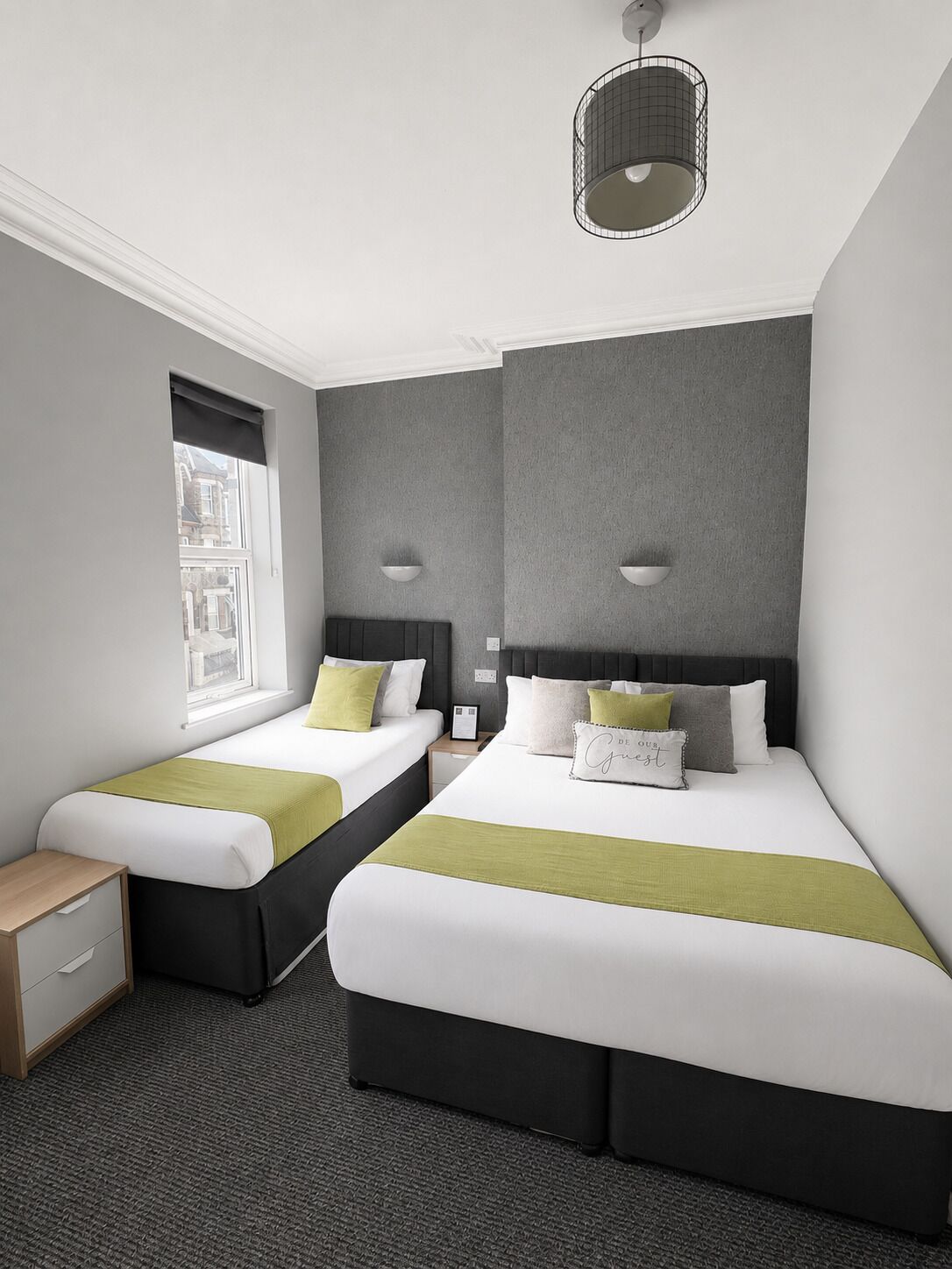 A hotel room with two beds, a window, a bedside table, and a hanging light fixture.