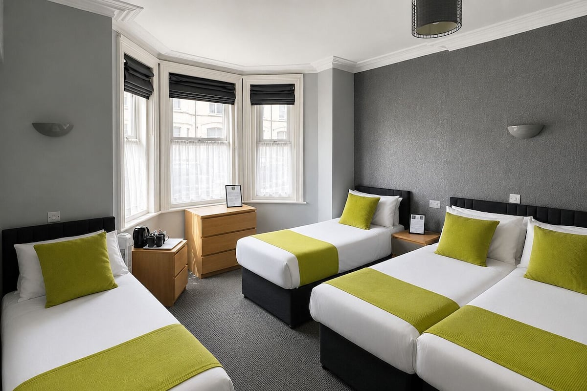 A hotel room with three beds, a wooden nightstand, a desk, and a window with blinds.