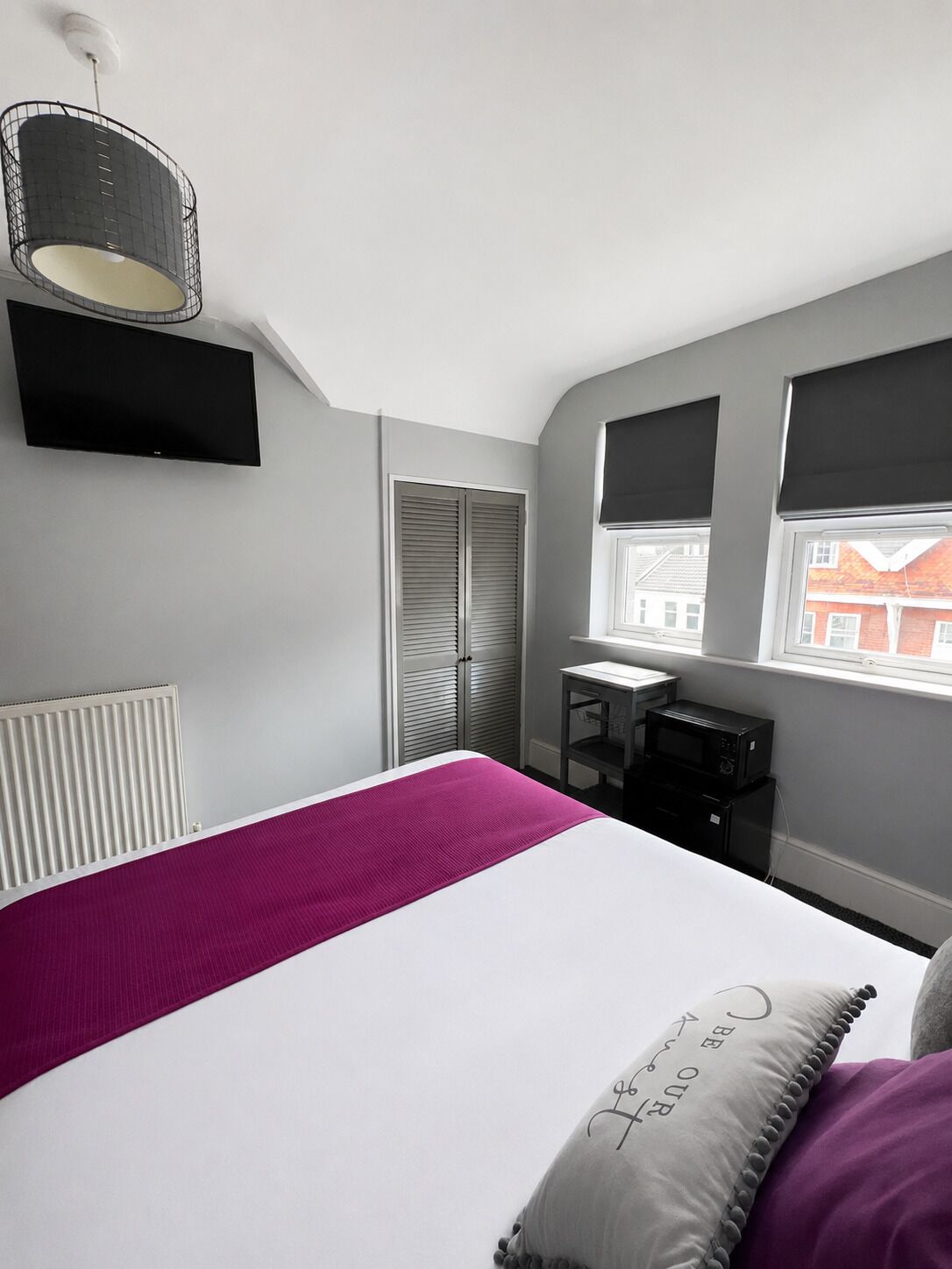 Un dormitorio con cama, escritorio, radiador, ventana y vistas a edificios.