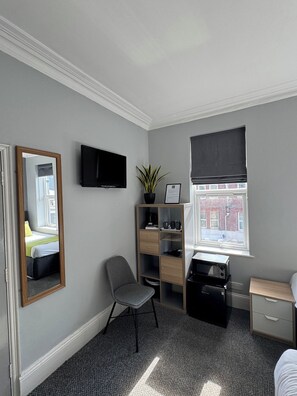A small, neatly arranged bedroom with a bed, a chair, a desk, a television, a window with a view, and a plant.