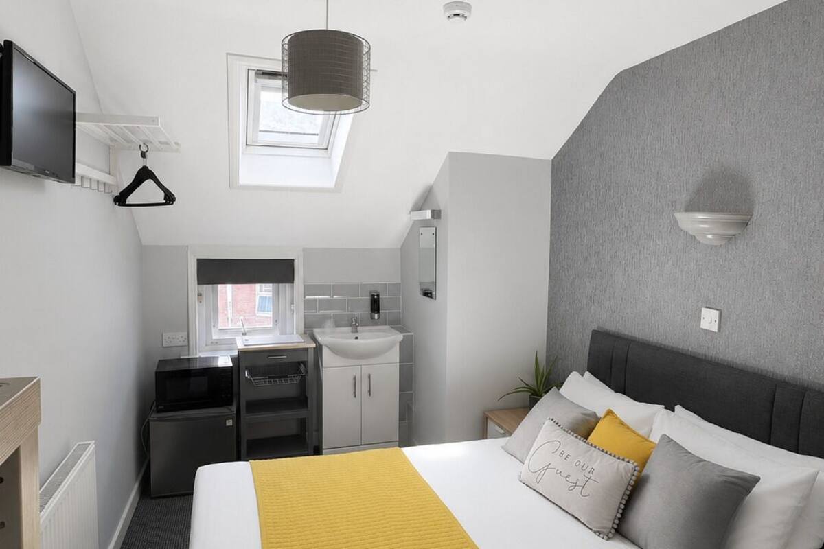 A compact hotel room with a bed, bedside table, desk, and a small kitchenette.