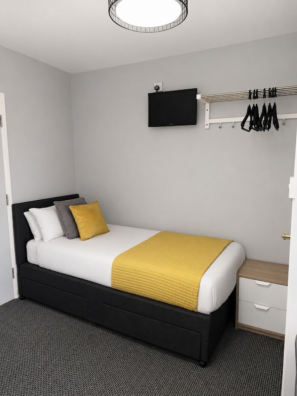 A modern bedroom with a black bed, white bedding, a yellow throw, a flat-screen TV, a coat rack, and a small bedside table.