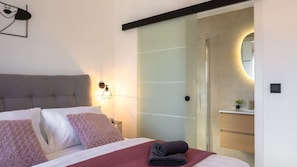Ein modernes Hotelzimmer mit einem Bett, einem Nachttisch, einer Wandlampe und einer Schiebetür zu einem Badezimmer.