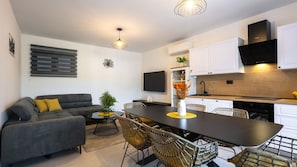 Eine moderne Küche mit weißen Schränken, eine Essecke mit dunklem Tisch und ein graues Sofa mit gelben Kissen.