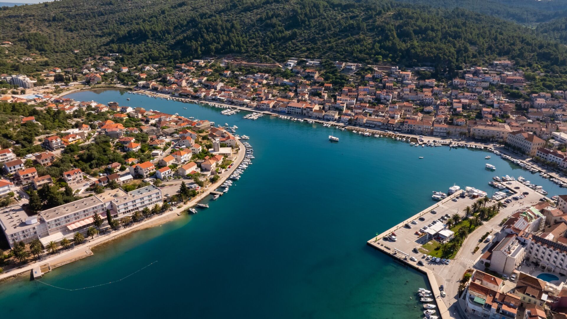 An aerial view of a coastal town with a marina, dense housing, and a mountainous backdrop.