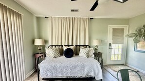 Ein Schlafzimmer mit einem Bett, zwei Nachttischen, einem Deckenventilator, einem Fenster mit Vorhängen und einer Tür.