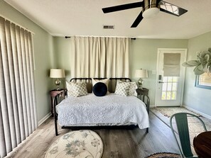 Ein Schlafzimmer mit einem Bett, zwei Nachttischen, einem Deckenventilator, einem Fenster mit Vorhängen und einer Tür.