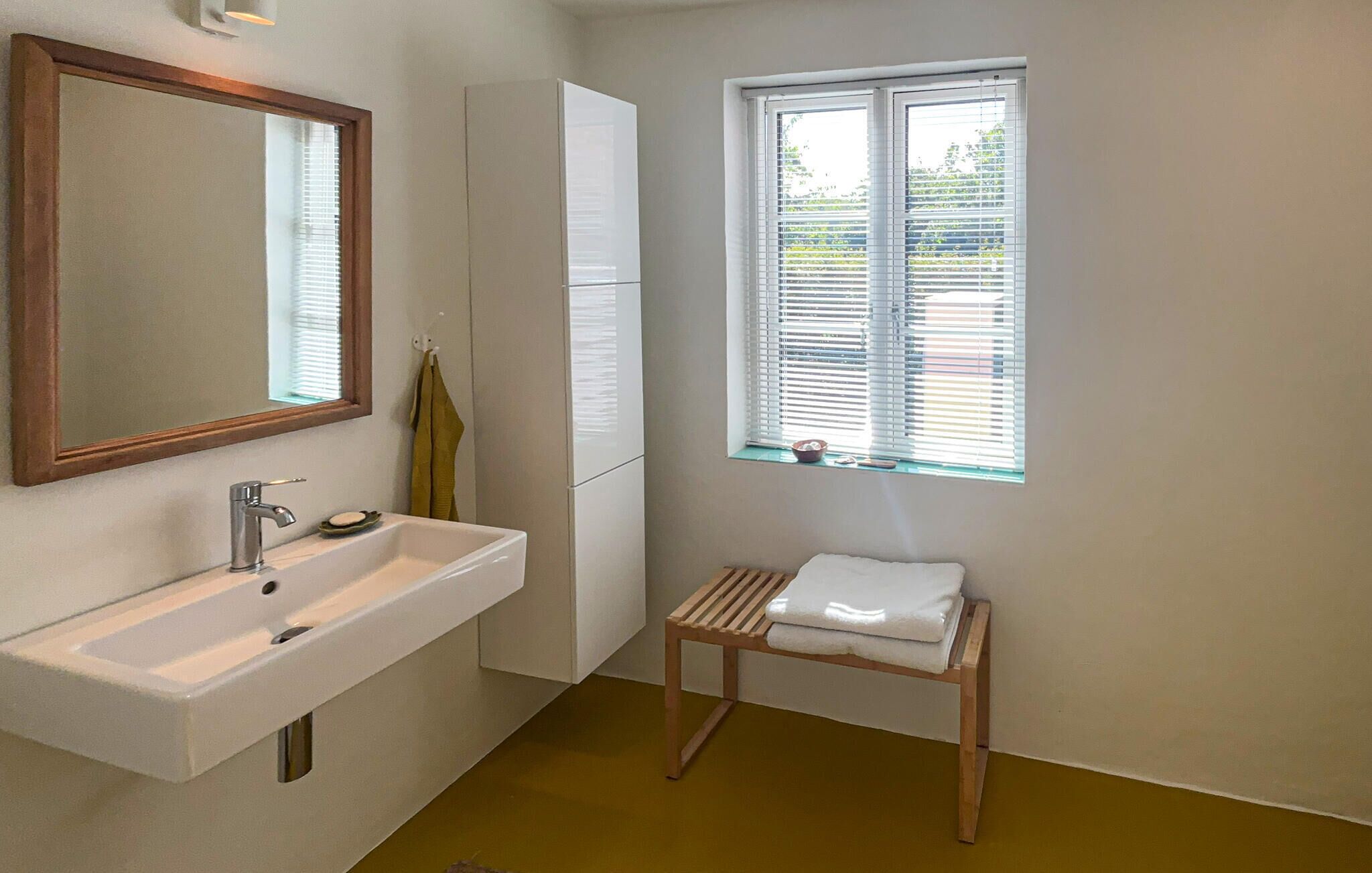 A bathroom with a white sink, a mirror, a window with blinds, and a wooden bench.