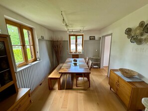 Uma sala de jantar com mesa de madeira, cadeiras, radiador, janela com vista e um ornamento na parede.