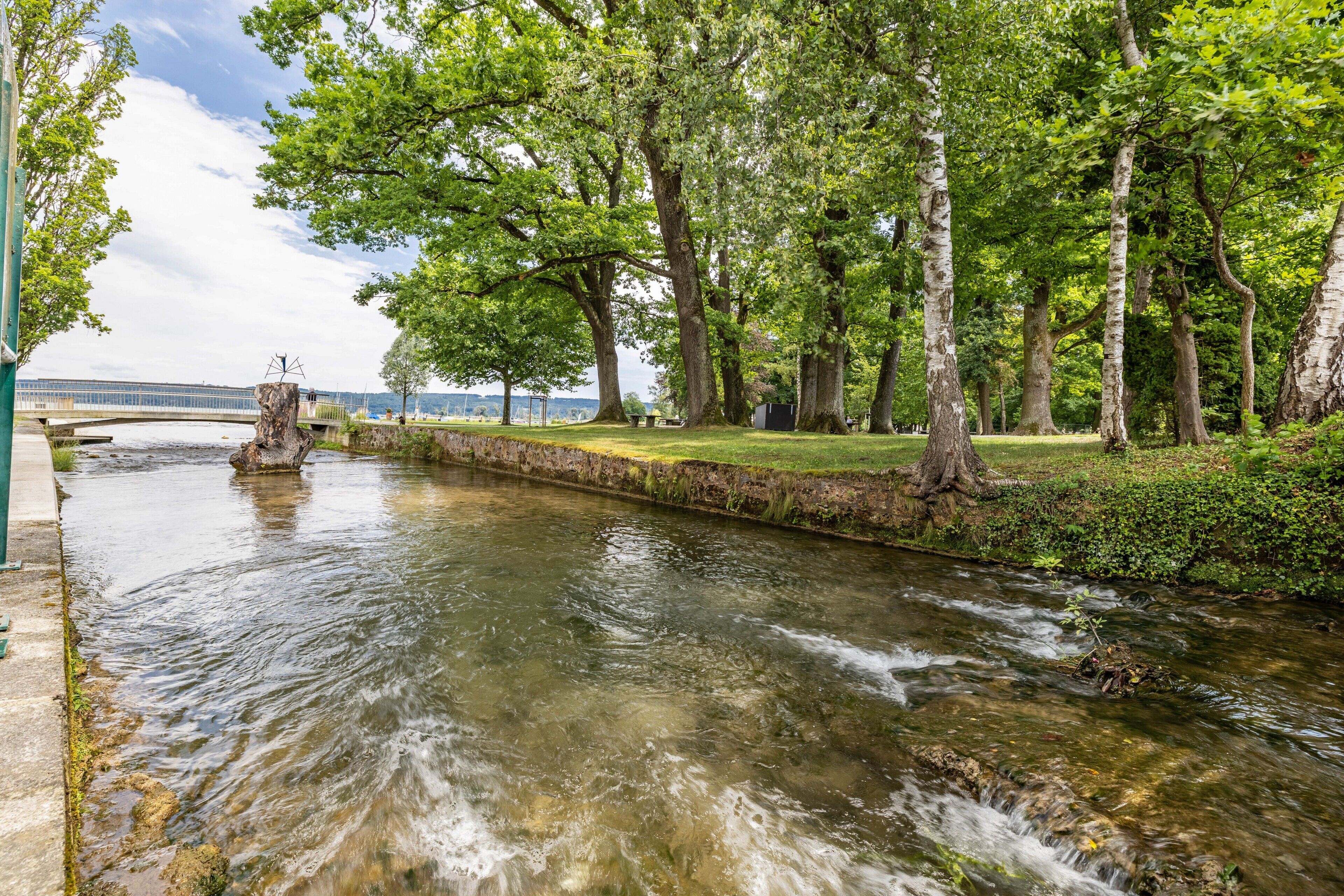 Ein ruhiger Fluss mit Bäumen am Ufer, eine Steinmauer und eine Metallskulptur.