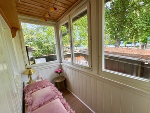A room with a wooden ceiling, a bench with pink cushions, a small table with a lamp, and a window with a view of trees and a fence. - Holiday Home 'Schick Am See' with Lake View, Private Terrace and Wi-Fi (Dießen am Ammersee)