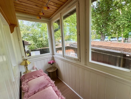 A room with a wooden ceiling, a bench with pink cushions, a small table with a lamp, and a window with a view of trees and a fence.. Holiday Home 'Schick Am See' with Lake View, Private Terrace and Wi-Fi