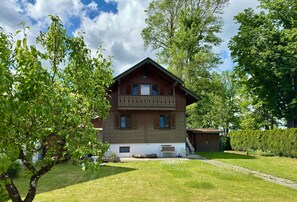 Uma casa de madeira com dois andares e varanda, rodeada por um gramado bem cuidado e árvores.